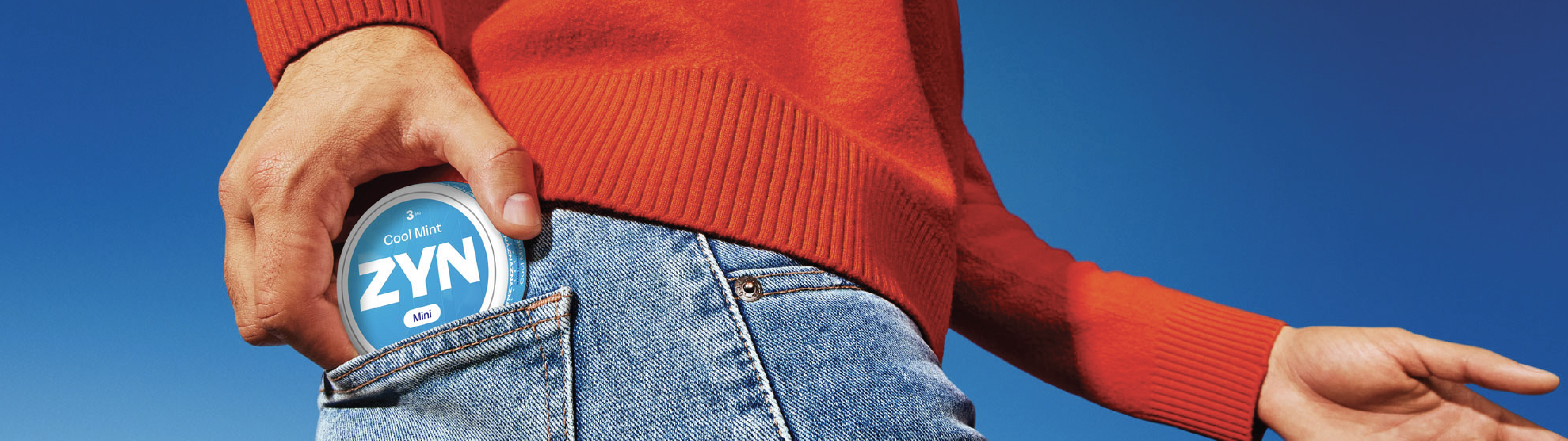 A person in a red sweater placing a red ZYN container into the back pocket of their blue jeans.
